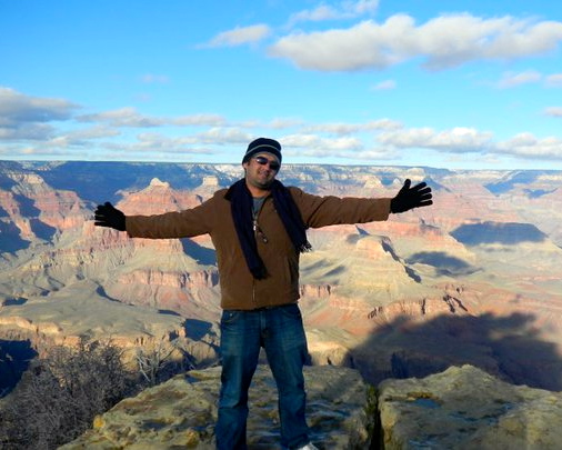 Abrash wearing a jacket, hat, gloves, and a scarf, standing with his hands our. He is standing at a peak at the Grand Canyon which is visible behind him.