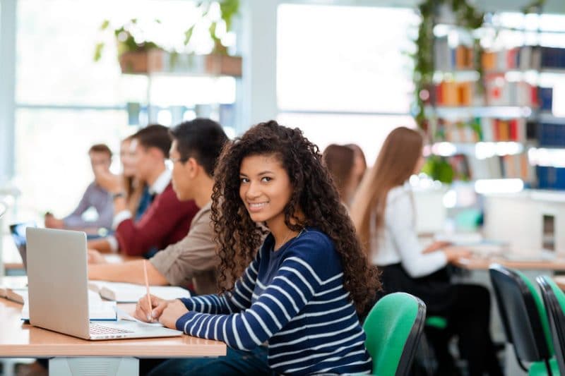 Attractive female student with long dark wavey hair smiling next to her laptop computer while taking notes in a library.