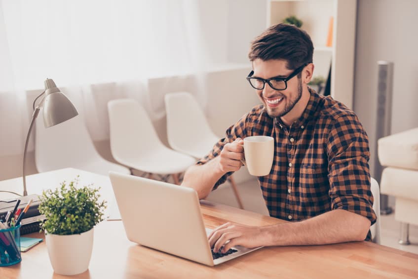 Young man wearing glasses, holding a mug, and sitting in front of his computer looking over an article about Postdoctoral Creative Writing Fellowships