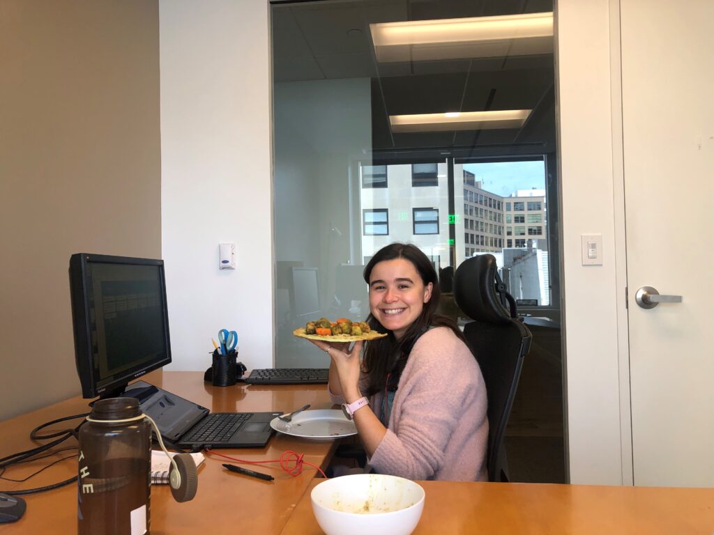 A young woman sitting at an office desk holding a plate of food and smiling.