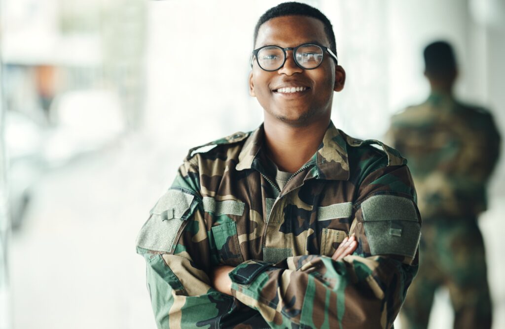 African American man, a military veteran, with his hands crossed, wearing camouflage, black glasses, and smiling, looking forward to applying to fellowships for veterans.