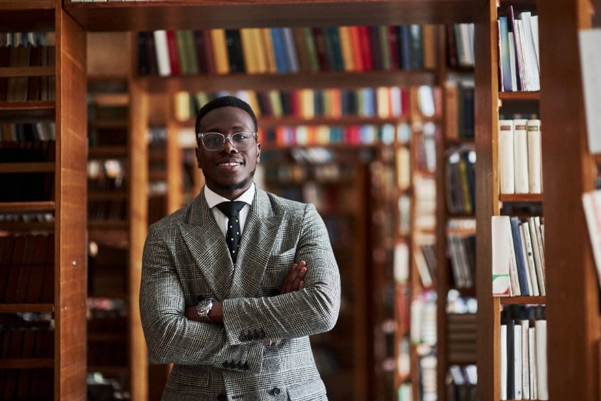 An African American man in a business suit smiling and standing with his arms folded in a library in the reading room