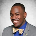 HBCUvc Fellow Nyland Sindifall smiling for a professional portrait wearing a grey suit, blue shirt, and a gold and white striped bow tie.