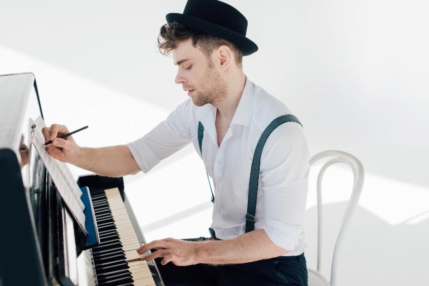 Focused male composer writing in his music book and playing a piano.