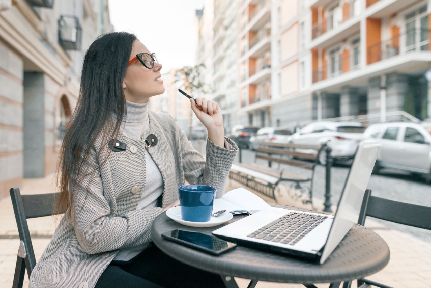 Young caucasian female student wearing glasses and a warm coat and sitting in an outdoor cafe in a city with a laptop computer and a cup of hot coffee and holding a pen while writing in her notebook.