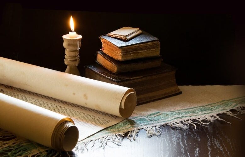 A scroll and old literary works piled on a dark wooden desk beside a burning candle.