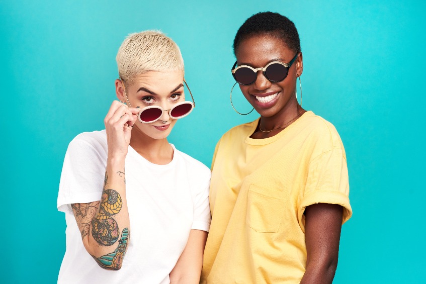 Two young women, one caucasian, one African-American, wearing sunglasses and smiling against a turquoise blue background.