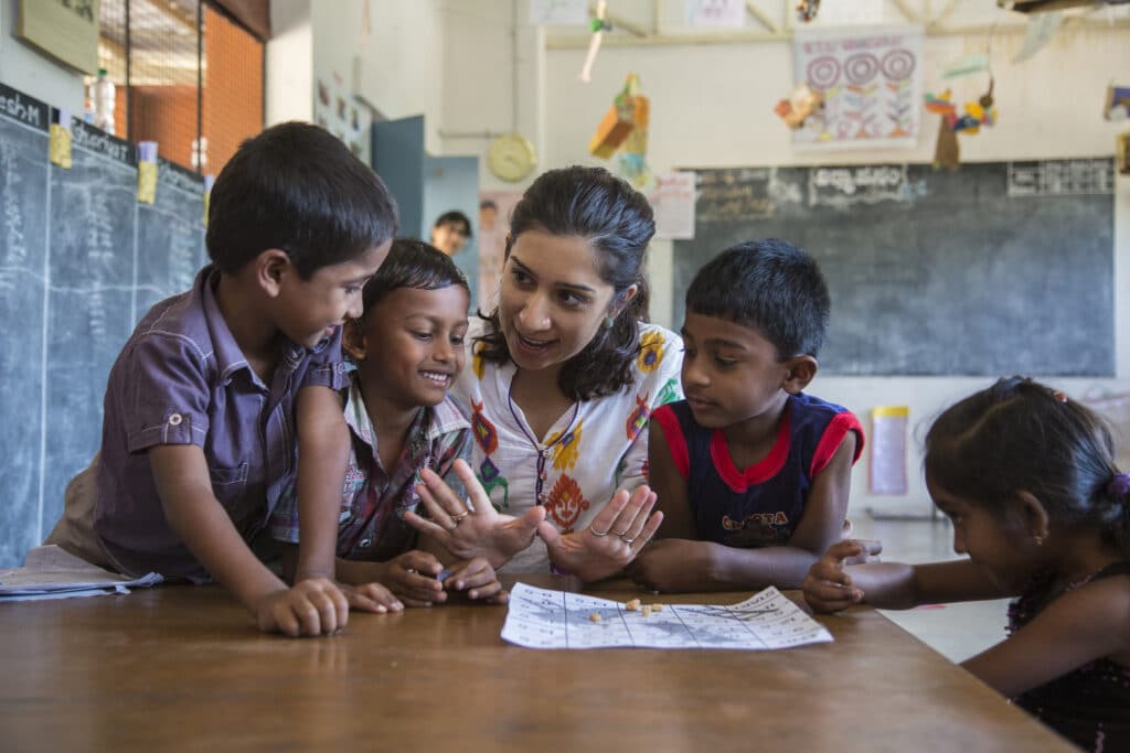 A Banyan Impact Fellow speaks to a group of four young children at a table in a schoolhouse.