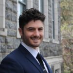 Nicholas Mancini headshot smiling in front of grey brick building. 