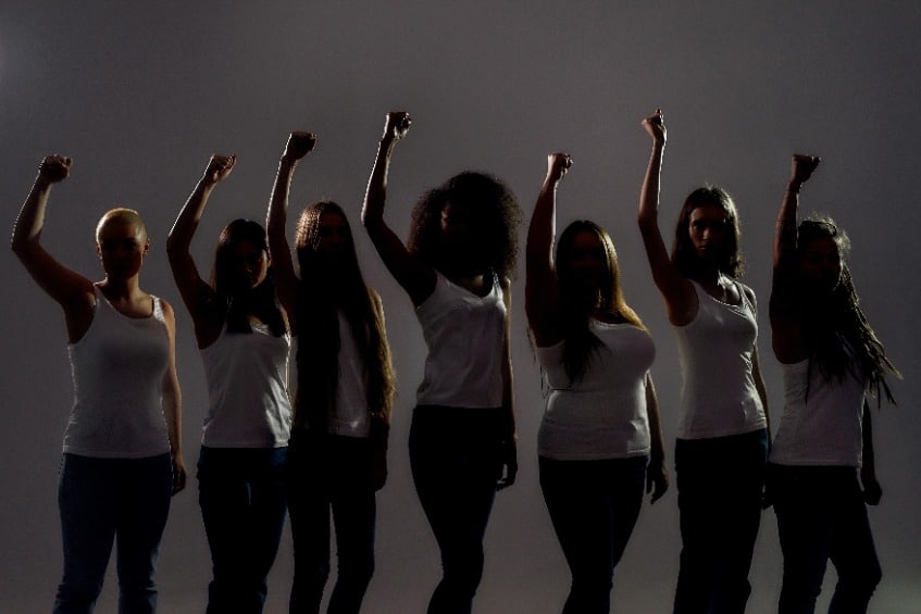 Silhouetted group of diverse women raised their arms, fists while standing over grey background.