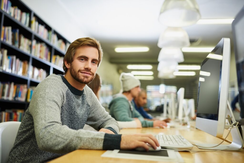 Young caucasian male university student wit ha beard and a grey sweater studying in the library.