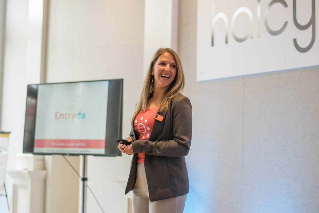 Erin Janklow in pink shirt, grey blazer, and beige pants standing in front of large computer screen giving presentation