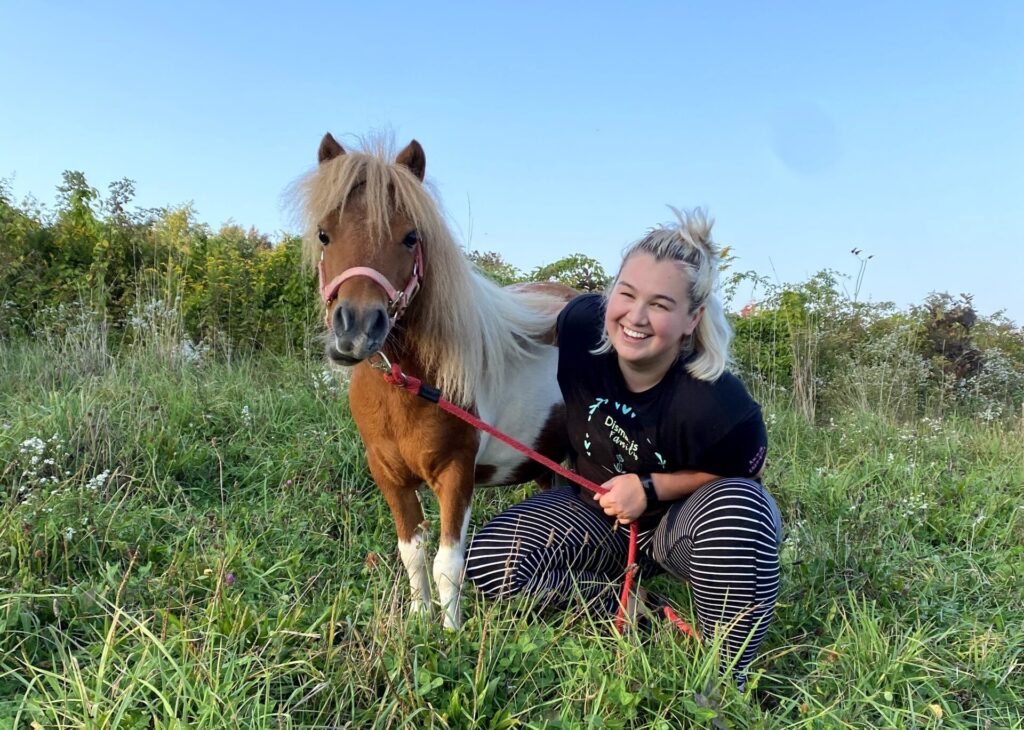 Dismas Family Farm Fellow Anna McCormack smiling in grassy field with a chestnut brown miniature horse