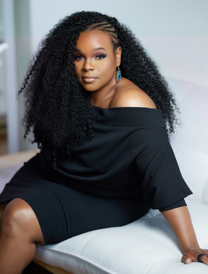 Kresge Arts Fellow Kisma Jordan-Hunter wearing a black dress and blue earnings and sitting on a couch.