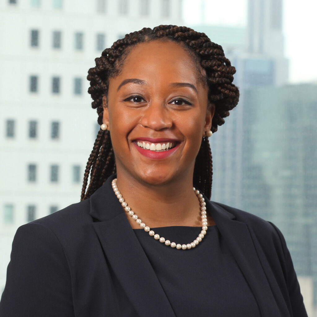 Ashlie Tyler smiling in a professional photo wearing a black jacket and pearls with a downtown scene in the background