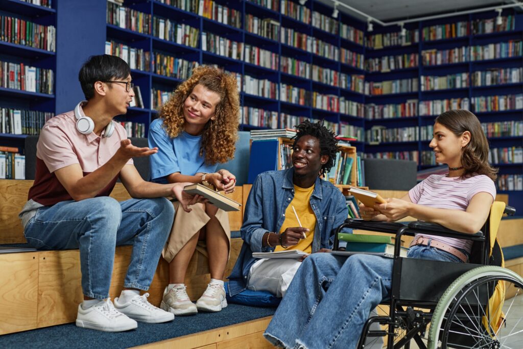 Diverse group of students in college library including young disabled woman in wheelchair enjoying discussion