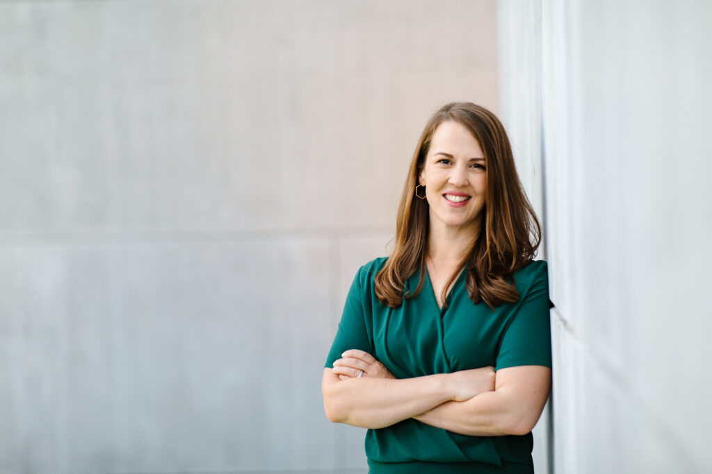 Dr. Vicki Johnson, founder of ProFellow™, smiling and wearing a green blouse with her arms folded and leaning up against a wall at a university.