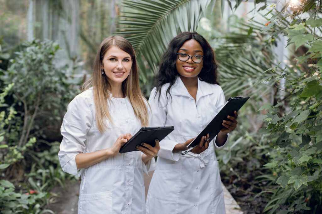 Two young pretty multiracial women botanists scientists, working in greenhouse with tropical plants and palm trees, smiling to camera with clipboard and tablet.