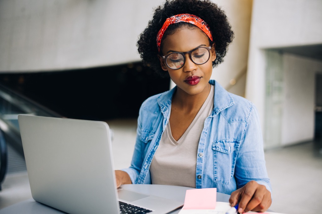 Confident young African female sitting at a table in an office studying for the GRE using a laptop and reading notes in her planner.