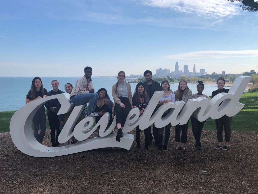 12 diverse young professionals representing the Cleveland Foundation Public Service Fellowship stand with the "Cleveland" sign at Edgewater Beach in Cleveland, Ohio.