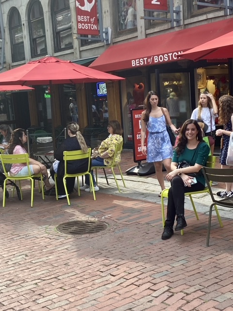 Dr. Vanessa Vieites seated at an outdoor table people watching near the Best of Boston gift shop on Market Street in Boston, Massachusetts, while on her AAAS Mass Media Science and Engineering Fellowship.