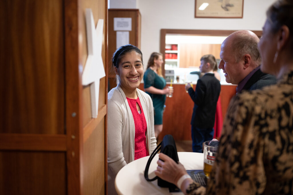 Fulbright ETA Magaly Cabrera-Ortiz in a red dress and light brown cardigan smiling for a picture while participating in a group discussion at a reunion with her school mentor and her husband to celebrate fourth-year students at Maturita Ball, before their final exams and graduation.