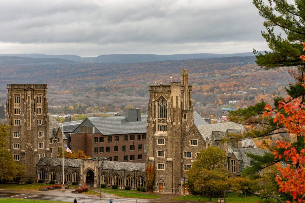 Cornell University in Ithaca, New York offers a Fully Funded PhD in Agricultural Sciences. Pictured are gothic stone buildings perched on a hill at Cornell University overlooking the tree covered valley below during the Autumn season with peak autumn leaf colors, including reds, oranges, and bright yellows.