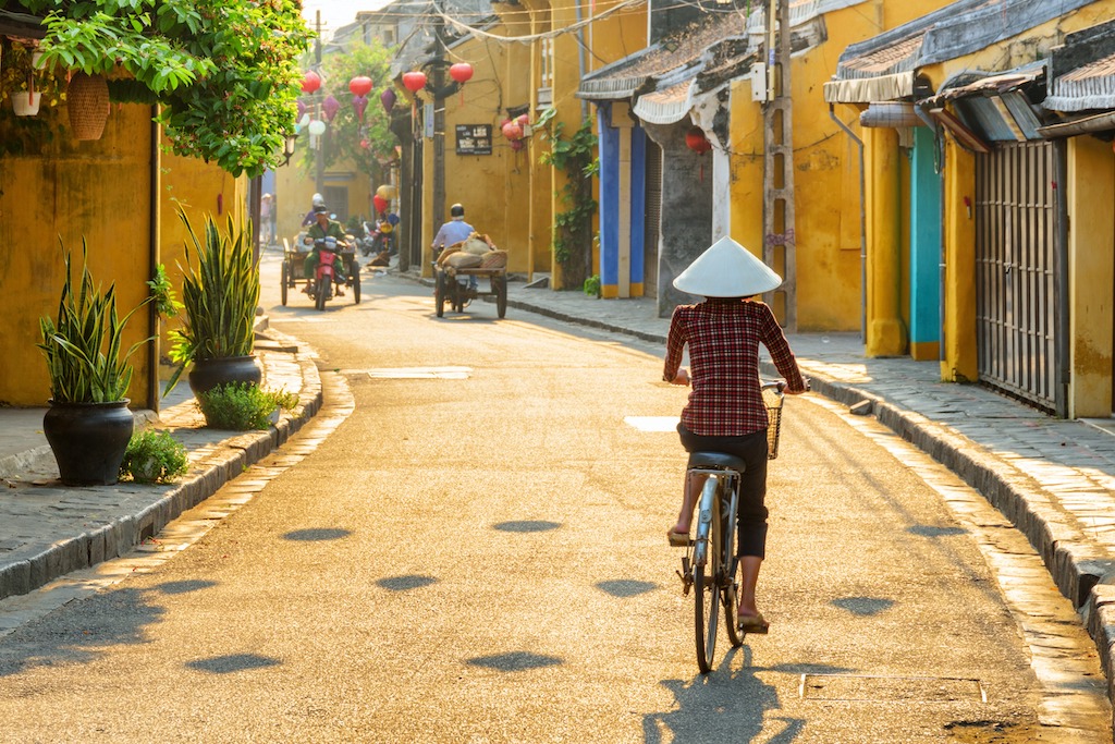In 2024-2025 Vietnam is offering the Fulbright-Fogarty Public Health Fellowship Award as part of the Fulbright U.S. Student Program. This is a picture of a Vietnamese woman in a traditional bamboo hat bicycling down a beautiful ancient street decorated with colorful silk lanterns at sunrise in Hoi An (Hon), Vietnam.