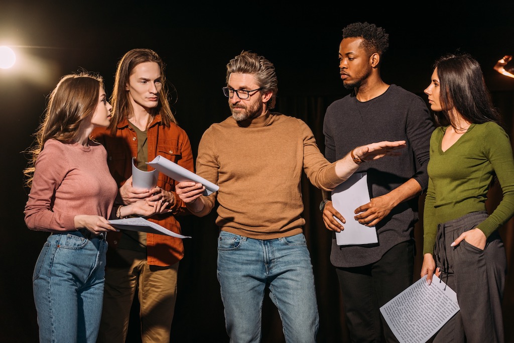 Theater fellowships provide funding and support for emerging and established theater professionals to hone their craft. In this picture there are multiethnic actors and actresses rehearsing with the theater director on stage.