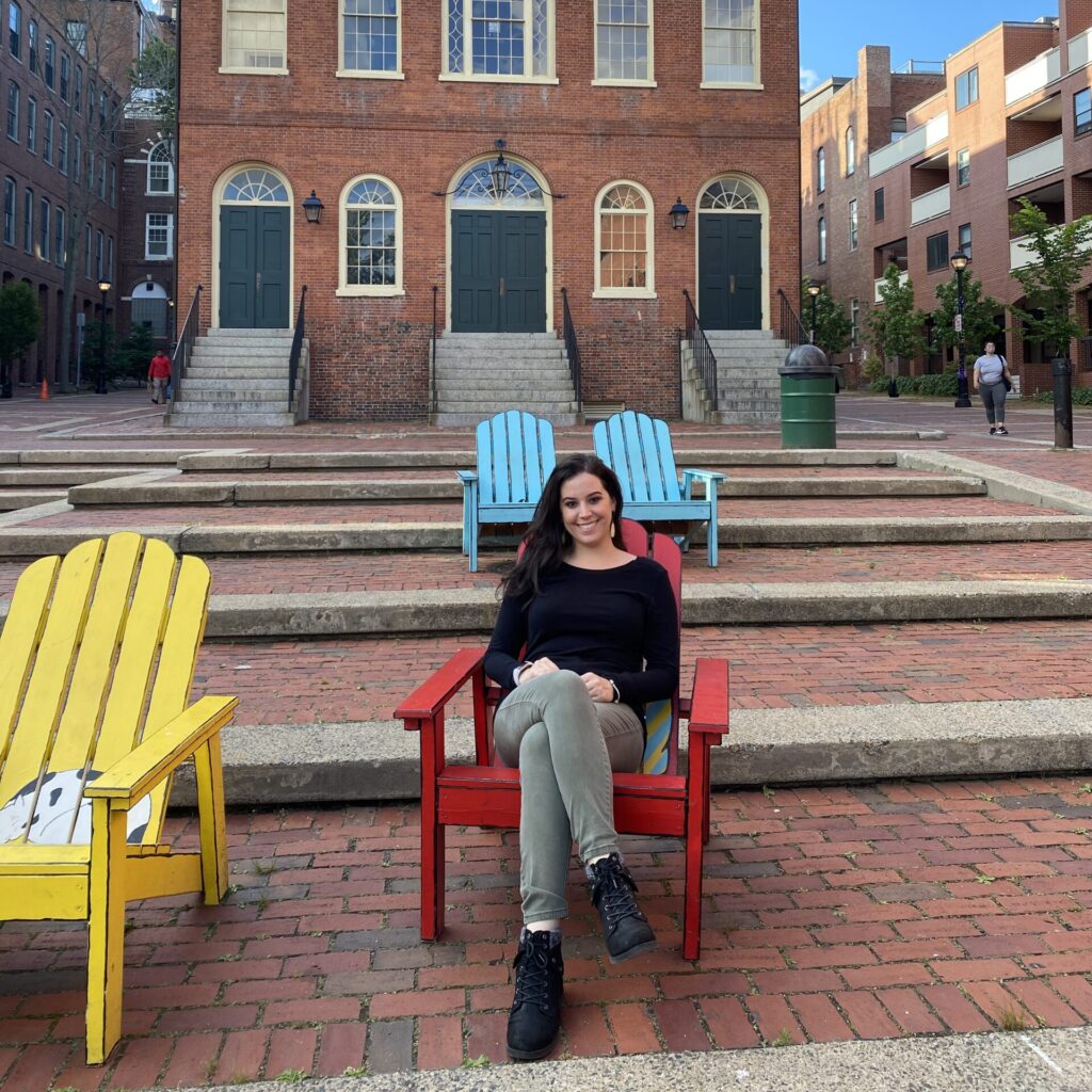Dr. Vanessa Vieites smiling while seated in a red Adirondack chair in front of the Town Hall building in Salem, Massachusetts, while on her AAAS Mass Media Science and Engineering Fellowship.