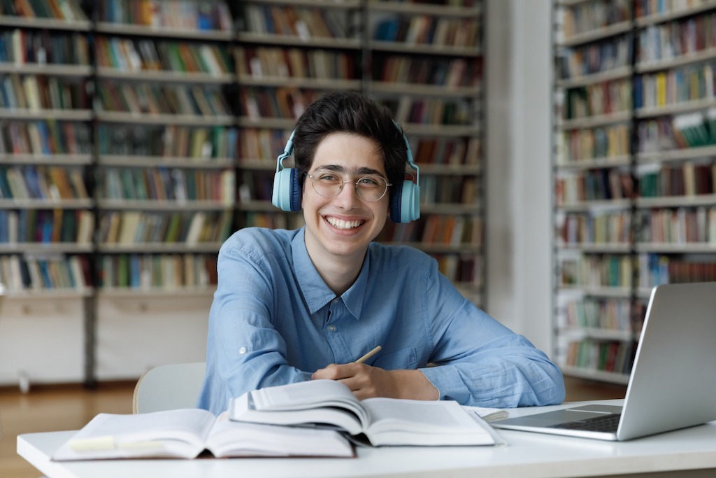 Smiling motivated male young Jewish leader working on applications for summer fellowships and professional fellowships that offer leadership training and Jewish studies for college students. He is in the library using his laptop computer to perform research while wearing headphones and taking notes in his notebook.