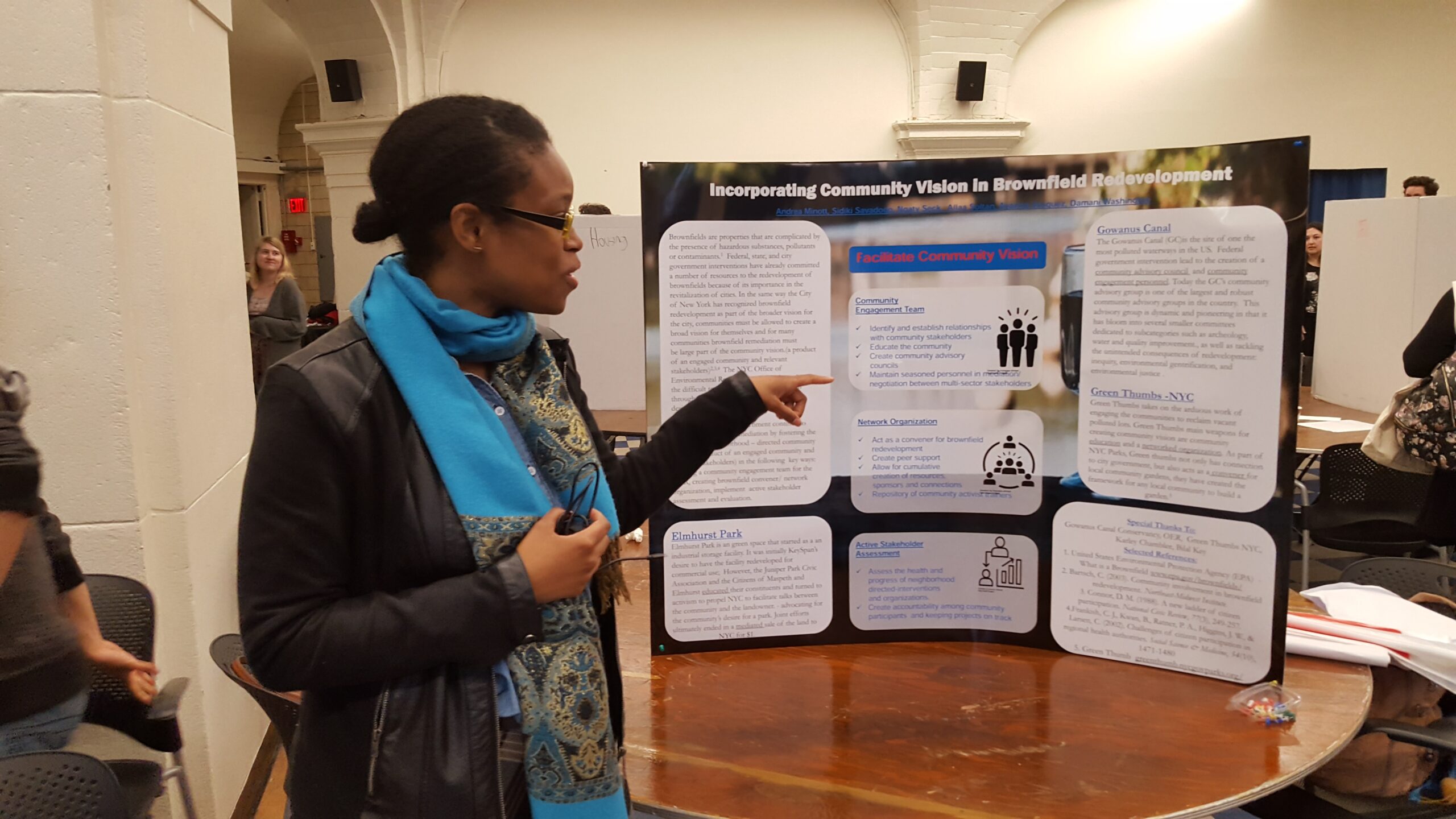 Ayanna Vasquez, fully funded MPH and preventative medicine residency at the University of Wisconsin-Madison, wearing a black jacket and bright blue scarf around her neck, stands by her poster board. With her hand stretched out and pointing, she presents a specific section of her her research into NYC brownfields.