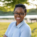 Professional head-shot of Ayanna Vasquez posing in front of lush green grass and a body of water in the background.