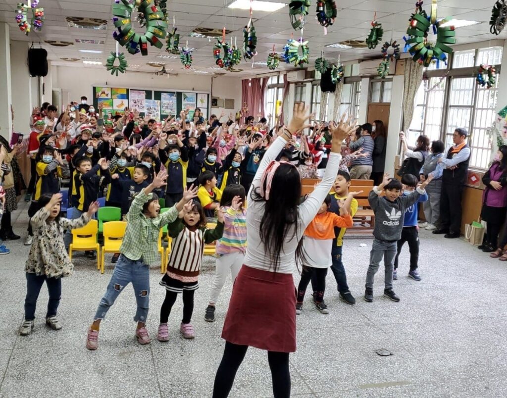 Fulbright ETA in Taiwan Sunny Huang leading the last dance for her school's Christmas concert. The classroom ceiling is decorated with Christmas decorations and dozens of students have their hands raised while dancing and singing in concert.