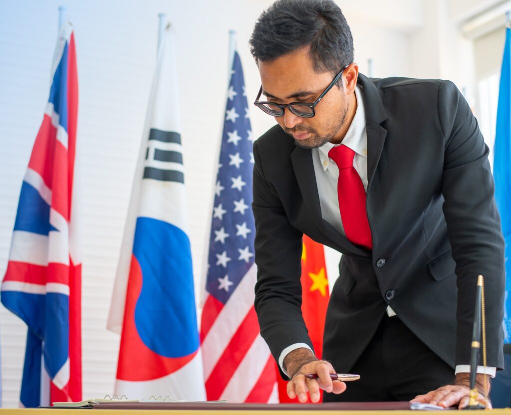 This list of universities that offer full funding to all or most students for PhD programs in Peace and Conflict Resolution. A man leans over a table with a pen in his hand. He is wearing glasses, a suite, white shirt, and red tie. Behind him are various country flags.