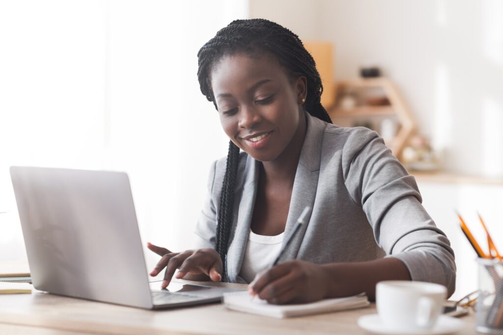 Young black female professional attending a fellowship training session for her remote fellowship. She is taking notes with a pen and paper while sitting in her home office and attending her fellowship meeting remotely.