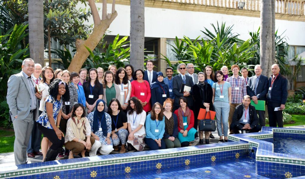 Nina Angeles (front row, right) with fellow Fulbright researchers at a conference in the MENA region. Three rows of students and mentors are standing and kneeling by the edge of a decorative pool. The sun is shining with palm trees in the background.