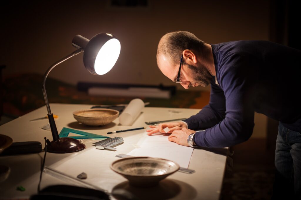 This list of upcoming fellowship deadlines includes a fully funded Master's program in Classics at the University of Kansas. In this image, a male archeologist examines ancient crocks at a university office under a lamp on a desk.