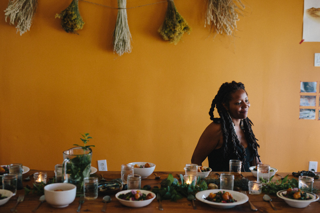 Dr. Mi'Jan Tho-Biaz sitting on the far right-hand side of a farm table with bowls and plates of food. There is a green, leafy garland on the table and glass jars with candles. The wall behind her is painted an orange color.