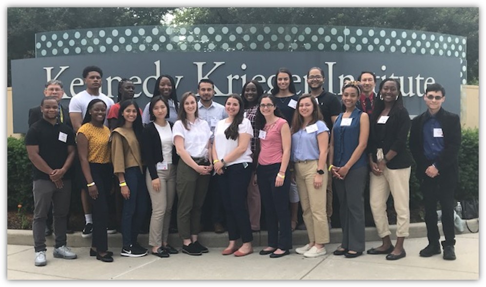 Previous years of Ferguson RISE fellowship winners posing in front of a large sign of the Kennedy Krieger Institute. The two rows of people consist of students and staff of various ethnicities.
