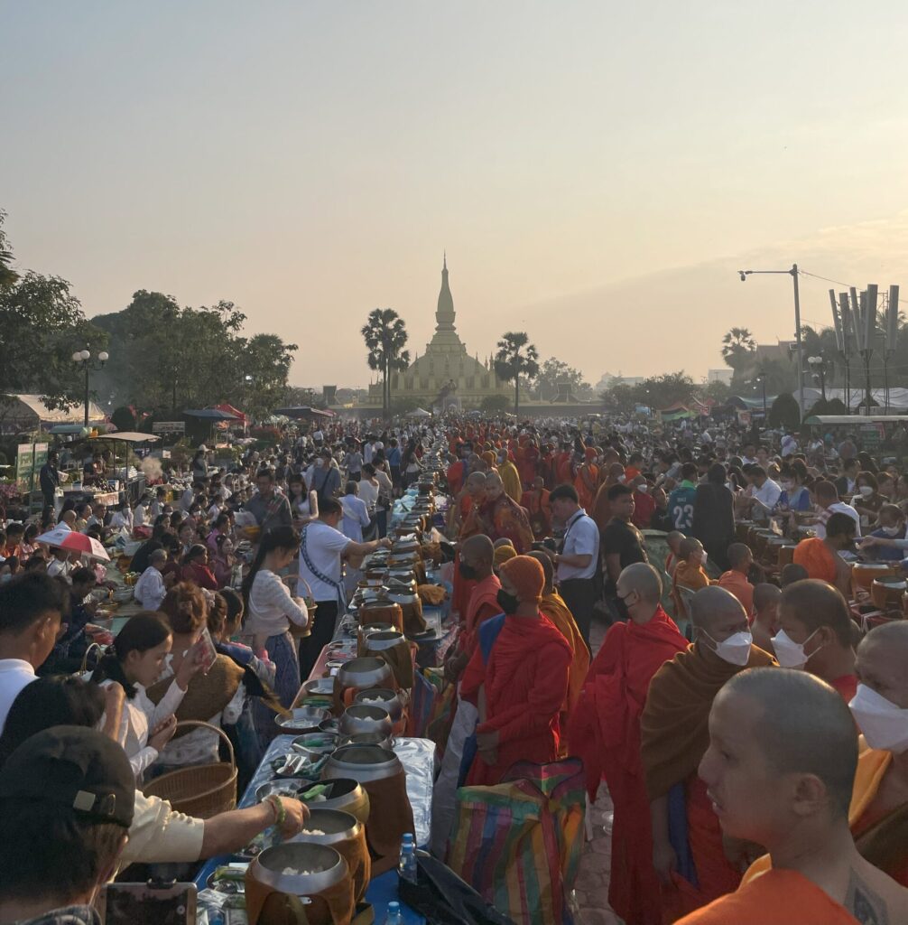 The That Luang Festival with Monks dressed in orange robes on the right-hand side sharing a feast with locals who take food from silver pots. Some people are praying, sitting on blankets, and wearing masks. The large crowd fills the entire picture with a temple that can be seen in the background.