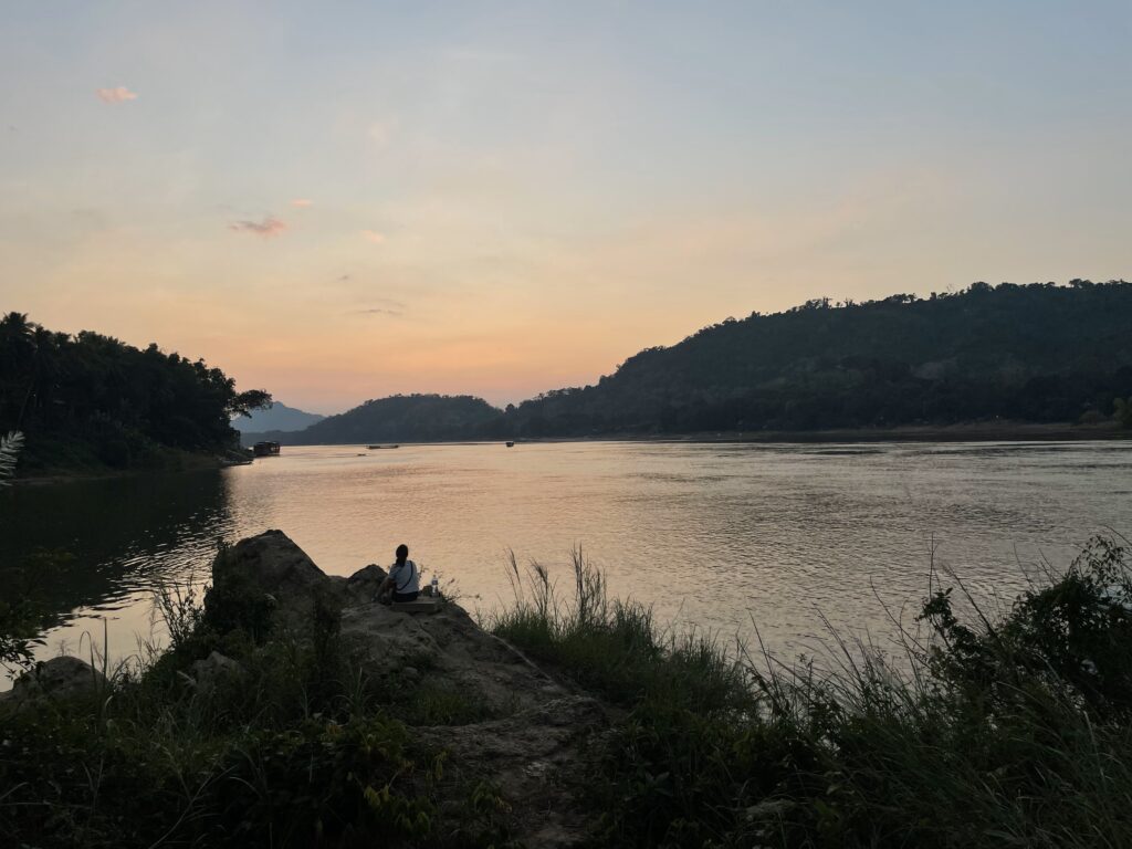 Maya sitting at the edge of a small hill by a river. The sun is setting and the river is a light shade of orange with the silhouette of Maya and river grass just visible.