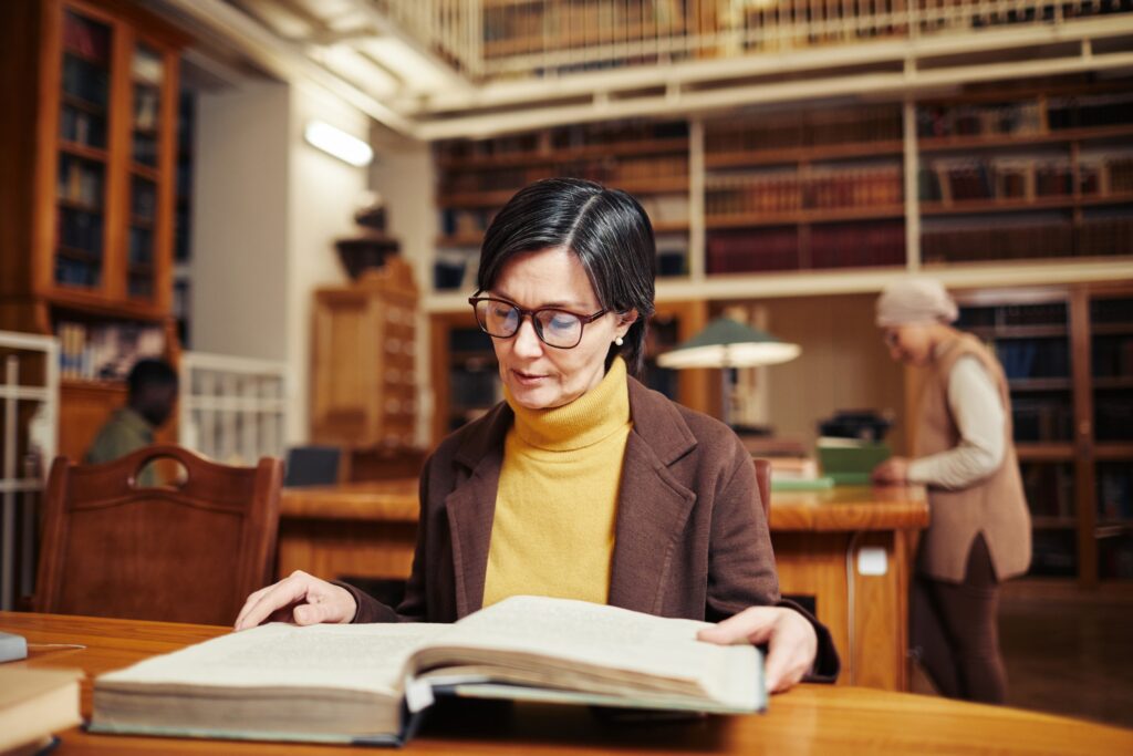 Older woman reading a book at a table in a research library.