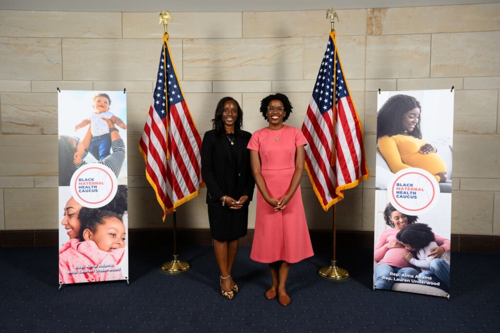 Lindsey Harris, DNP, who was awarded the RWJF Health Policy Fellowship, is wearing a black dress and suit jacket and standing next to Representative Lauren Underwood of Illinois, wearing an orange-pink short-sleeved dress. They are at the Maternal Health Caucus 2023 Stakeholder Summit with American Flags and summit promotional banners on either side.