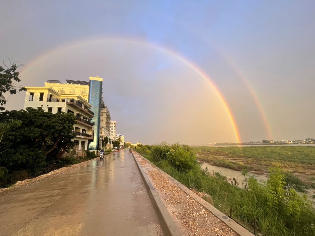 After heavy rain, a double rainbow appears over the Mekong River in Vientiane, the capital city of Laos. There is a pathway in the center, with a building to the left and the river on the right-hand side, with the rainbow fully encompassing both. This picture was taken by Princeton in Asia Fellowship winner Maya Bian.