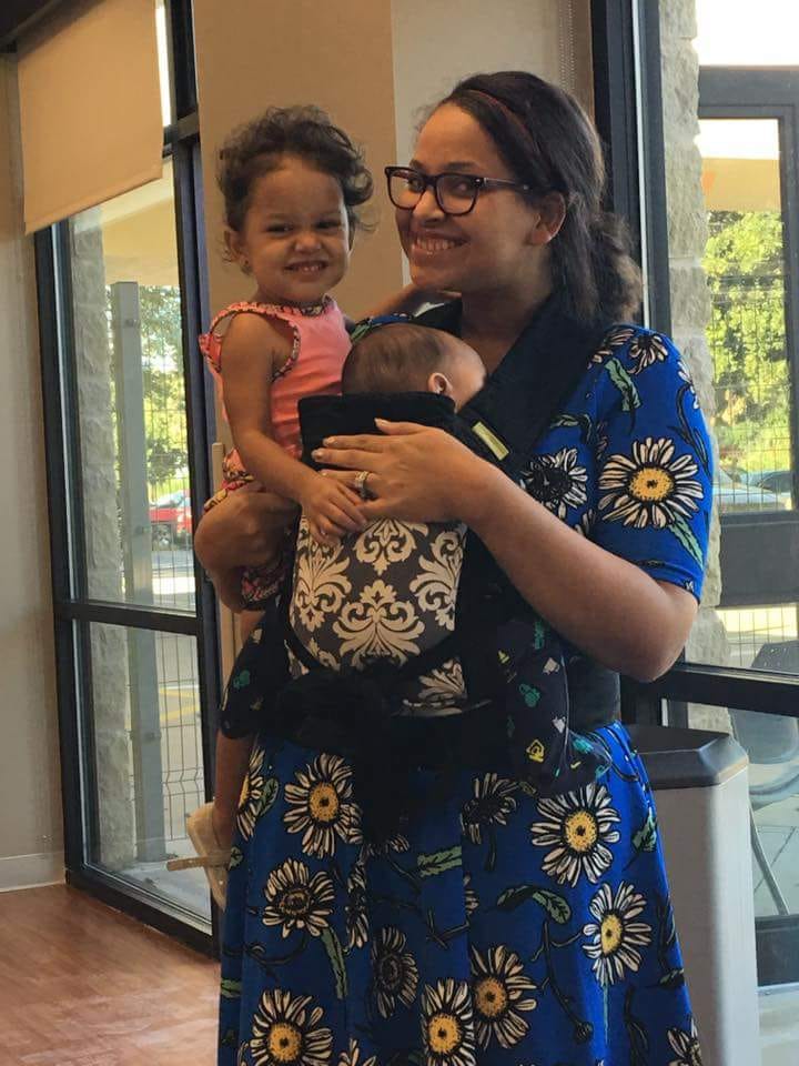 Global Health Corps Fellow Paris Acosta, wearing a dark blue dress with yellow flowers, holding her daughter Khloe, and her infant son Cameron, in a baby carrier.