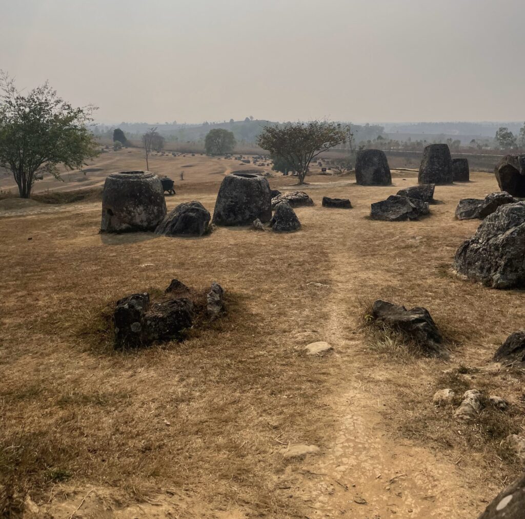 The Plain of Jars Site 1 in Xieng Khouang Province which has large pot-like structures scattered across dry. brown grass. Some craters can be seen, indicative of old bombs that have gone off. This picture was taken by Princeton in Asia Fellowship winner Maya Bian.