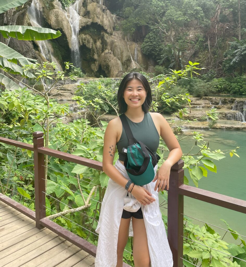 Maya Bian, Princeton in Asia Fellowship winner, standing on a wooden bridge in front of Kuangsi Waterfall in Luang Prabang, Laos. Behind her is lush greenery and the pools of water at the end of a waterfall.