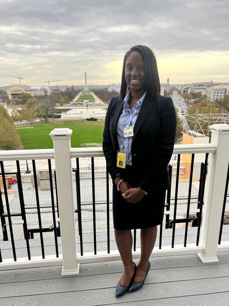 Lindsey Harris, smiling and standing at the Speaker of the House’s white and black balcony. She overlooks the Washington Monument in the background on a partly cloudy day.