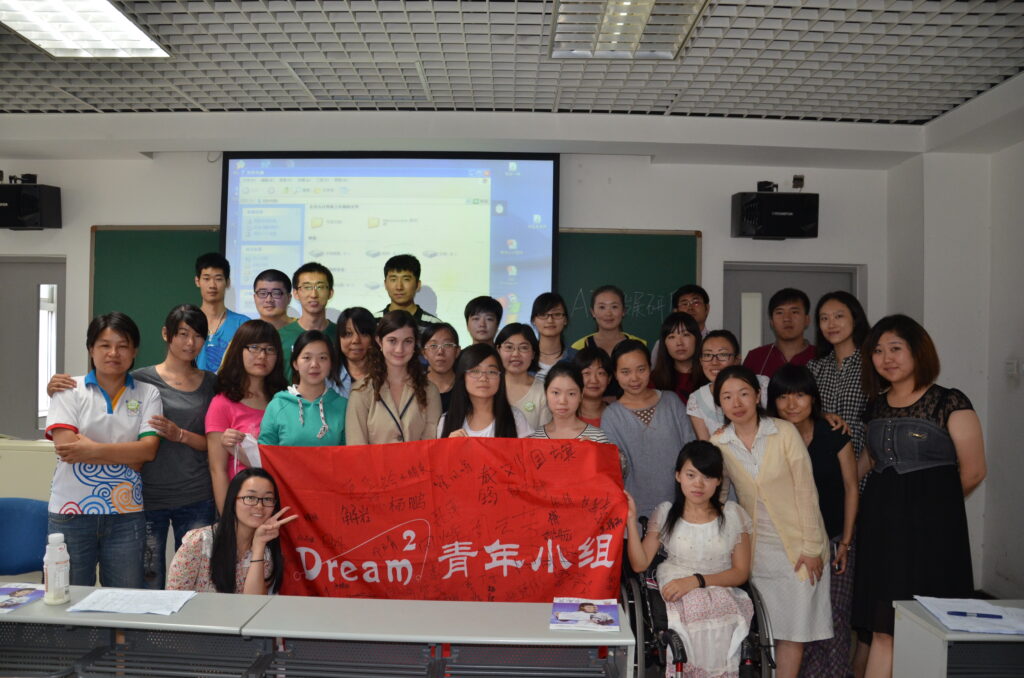 Multi Fellowship winner Meghan stands in a classroom full of Chinese students, with all of them holding up a red banner that reads "Dream squared Youth Group" in a mix of English and Mandarin